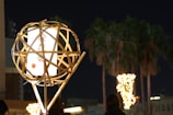 A bamboo lantern with a glowing white paper inside is situated in the foreground, featuring calligraphy and a small red circle. In the background, illuminated palm trees line an urban street at night.