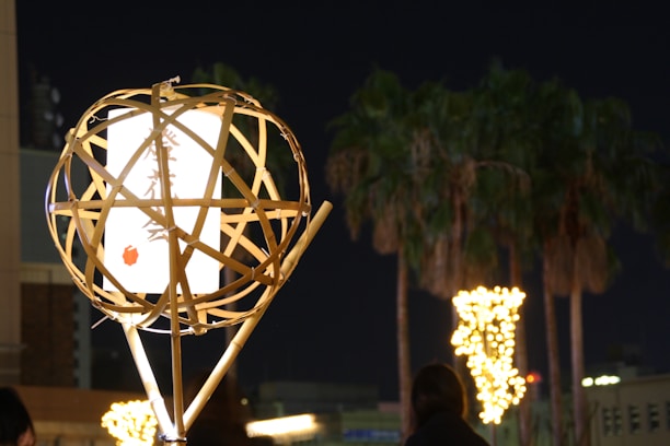 A bamboo lantern with a glowing white paper inside is situated in the foreground, featuring calligraphy and a small red circle. In the background, illuminated palm trees line an urban street at night.