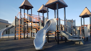 A playground structure composed of multiple slides, climbing ladders, and platforms with orange rails and gray surfaces. The equipment is set on a dark rubberized surface and surrounded by a building.