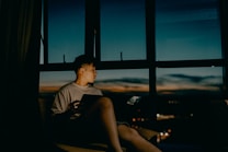 A person sits in front of a large window during twilight, looking outside at a serene evening sky. They hold a device, possibly a tablet, in their lap. The room is dimly lit, creating a calm and reflective atmosphere, contrasting with the deep blues and oranges of the sky.