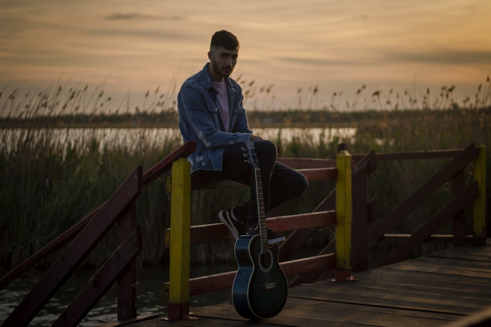 man in blue denim jacket and black pants sitting on brown wooden fence during daytime