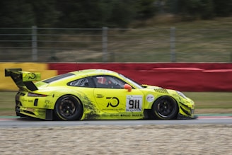 A bright yellow sports car with various sponsor logos races down a track. The car features a prominent rear spoiler and number 911 displayed on the side. The background includes a blurred red and yellow barrier and greenery, indicating high speed.