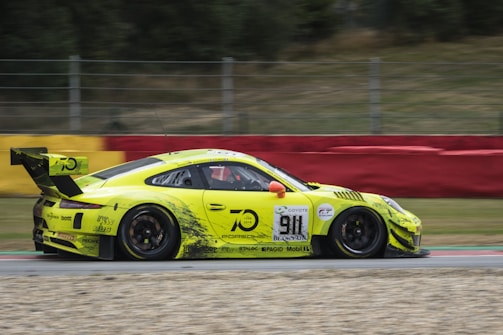 A bright yellow sports car with various sponsor logos races down a track. The car features a prominent rear spoiler and number 911 displayed on the side. The background includes a blurred red and yellow barrier and greenery, indicating high speed.