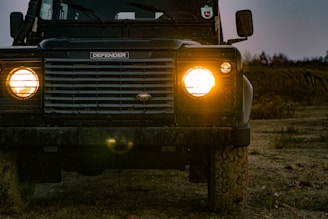 A rugged off-road vehicle is parked on a dirt path, with its headlights turned on, illuminating the surrounding area. The vehicle appears sturdy and designed for challenging terrains.