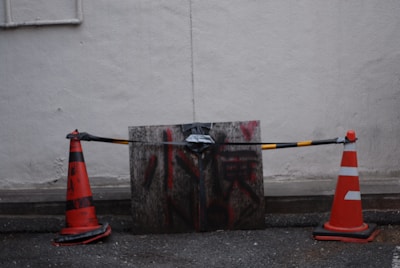 Two orange traffic cones are placed on either side of a wooden board that has been secured with a black and yellow striped tape. The board appears weathered with red and black graffiti. The setup is against a plain, textured gray wall, and the ground is a rough, asphalt surface.