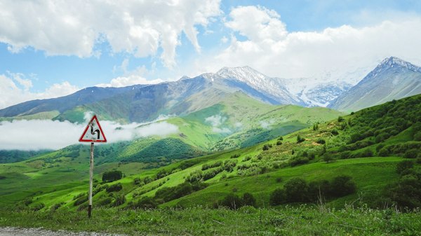 Green pastures below snow peaks on the road in — Red and white house on green grass field near mountains under white clouds and blue sky