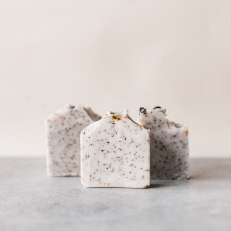 Three rectangular bars of handmade soap, speckled with small brown flecks, are placed on a neutral, light-colored surface. The soap has a natural, rustic appearance with some imperfections and irregularities on the top edges.