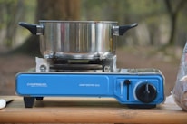 A camping stove with a blue base is set up on a wooden surface outdoors. A stainless steel pot with black handles is resting on the stove. The background is blurred with hints of greenery and trees.