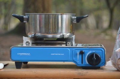 A camping stove with a blue base is set up on a wooden surface outdoors. A stainless steel pot with black handles is resting on the stove. The background is blurred with hints of greenery and trees.