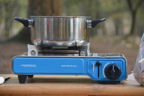 A camping stove with a blue base is set up on a wooden surface outdoors. A stainless steel pot with black handles is resting on the stove. The background is blurred with hints of greenery and trees.