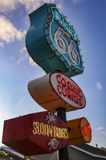 A vintage roadside sign featuring retro-style neon lights against a partly cloudy sky. The sign is composed of several sections displaying text such as 'Buck's on 66', 'Cosmic Curios', and 'Souvenirs', evoking nostalgia for historic Route 66.