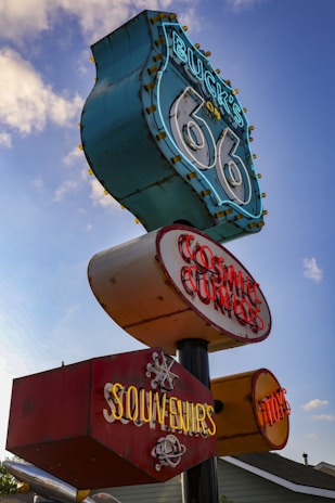 A vintage roadside sign featuring retro-style neon lights against a partly cloudy sky. The sign is composed of several sections displaying text such as 'Buck's on 66', 'Cosmic Curios', and 'Souvenirs', evoking nostalgia for historic Route 66.