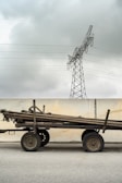 A cart stacked with long wooden poles stands in front of a concrete wall. Above, a metal power line tower looms against a cloudy sky.