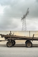 A cart stacked with long wooden poles stands in front of a concrete wall. Above, a metal power line tower looms against a cloudy sky.