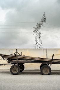 A cart stacked with long wooden poles stands in front of a concrete wall. Above, a metal power line tower looms against a cloudy sky.