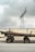 A cart stacked with long wooden poles stands in front of a concrete wall. Above, a metal power line tower looms against a cloudy sky.