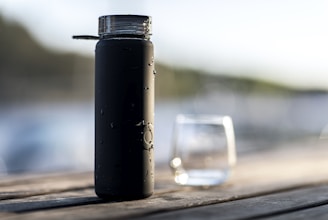 A matte black water bottle with condensation droplets on its surface stands on a wooden table. In the background, a blurred glass is visible along with a natural setting, suggesting an outdoor environment near water.