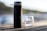 A matte black water bottle with condensation droplets on its surface stands on a wooden table. In the background, a blurred glass is visible along with a natural setting, suggesting an outdoor environment near water.