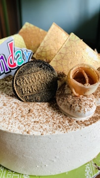 A close-up view of a decorative cake with creamy frosting, sprinkled lightly with cocoa powder. There are chocolate elements shaped like coins and geometric pieces, along with a scroll-like chocolate garnish. A colorful sign partially visible indicates a celebratory occasion.