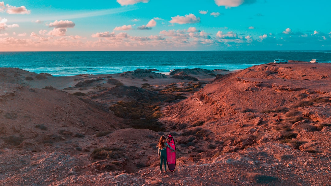 woman in blue jacket and blue denim jeans standing on brown rock formation near body of on with on with