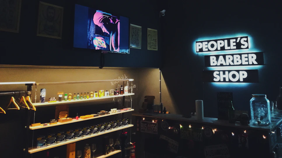 A cozy barbershop interior with a modern, dimly lit ambiance. Shelves are stocked with grooming products and a neon sign reading 'PEOPLE'S BARBER SHOP' is prominently displayed on a dark wall. A television screen shows an image of a person wearing a turban, adding to the shop's unique style.