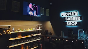 A cozy barbershop interior with a modern, dimly lit ambiance. Shelves are stocked with grooming products and a neon sign reading 'PEOPLE'S BARBER SHOP' is prominently displayed on a dark wall. A television screen shows an image of a person wearing a turban, adding to the shop's unique style.