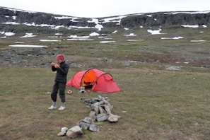 A person wearing a red hat and black jacket stands next to a small red tent in a grassy, mountainous area with patches of snow. The person appears to be eating, while camping equipment and a circle of stones are nearby. The landscape features rocky terrain and a backdrop of cliffs and patches of white snow.