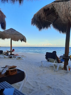 A tranquil beach scene with soft white sand and clear blue skies. Straw parasols provide shade for loungers scattered along the shore. The calm sea stretches to the horizon with gentle waves lapping at the beach. A few people are relaxing on sun loungers, enjoying the peaceful environment. Personal items like bags and towels are visible next to the loungers.