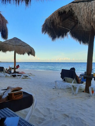 A tranquil beach scene with soft white sand and clear blue skies. Straw parasols provide shade for loungers scattered along the shore. The calm sea stretches to the horizon with gentle waves lapping at the beach. A few people are relaxing on sun loungers, enjoying the peaceful environment. Personal items like bags and towels are visible next to the loungers.