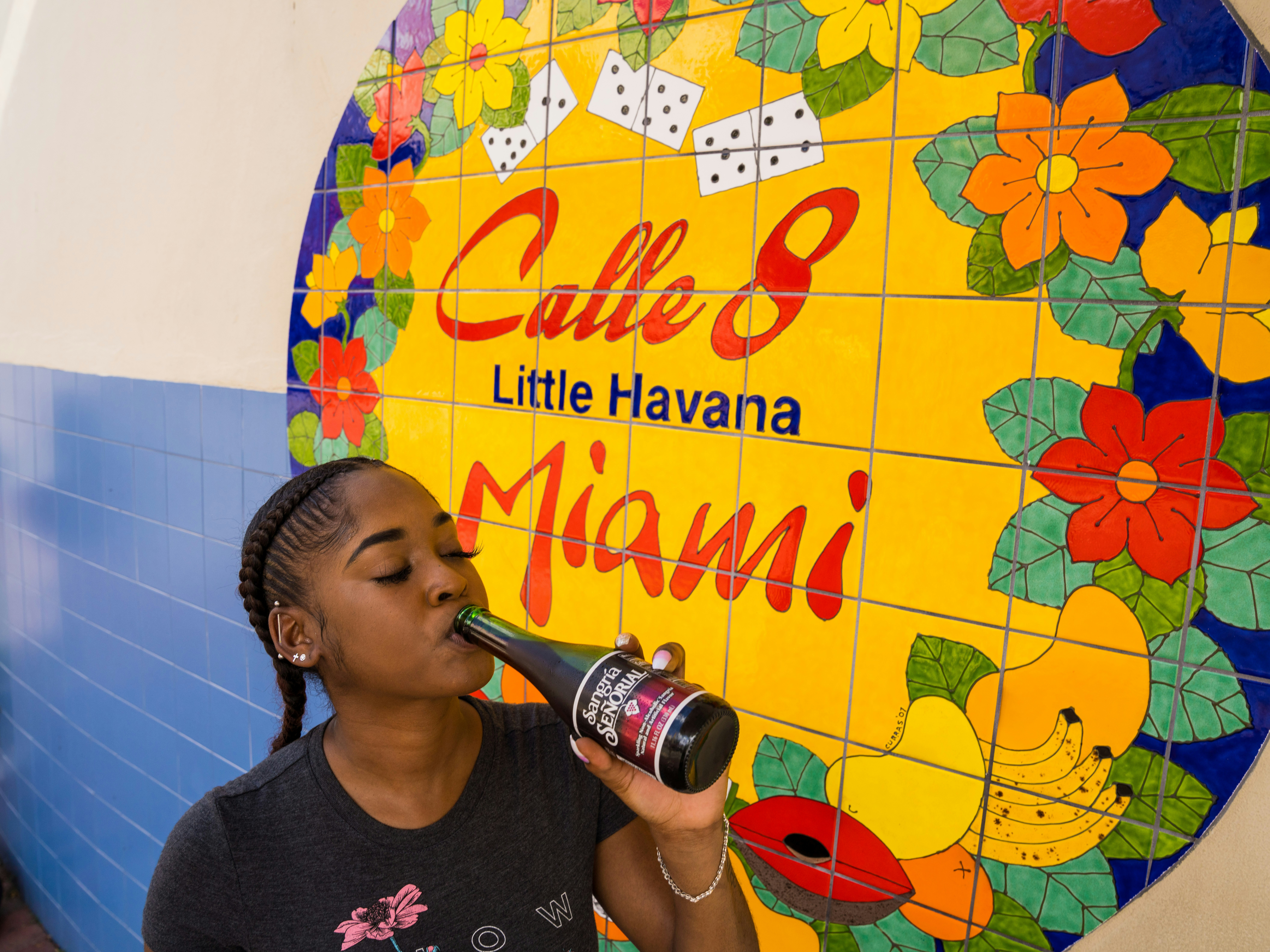 man in black crew neck t-shirt drinking from bottle , text at wall “Calle 8, Little Havana, Miami”