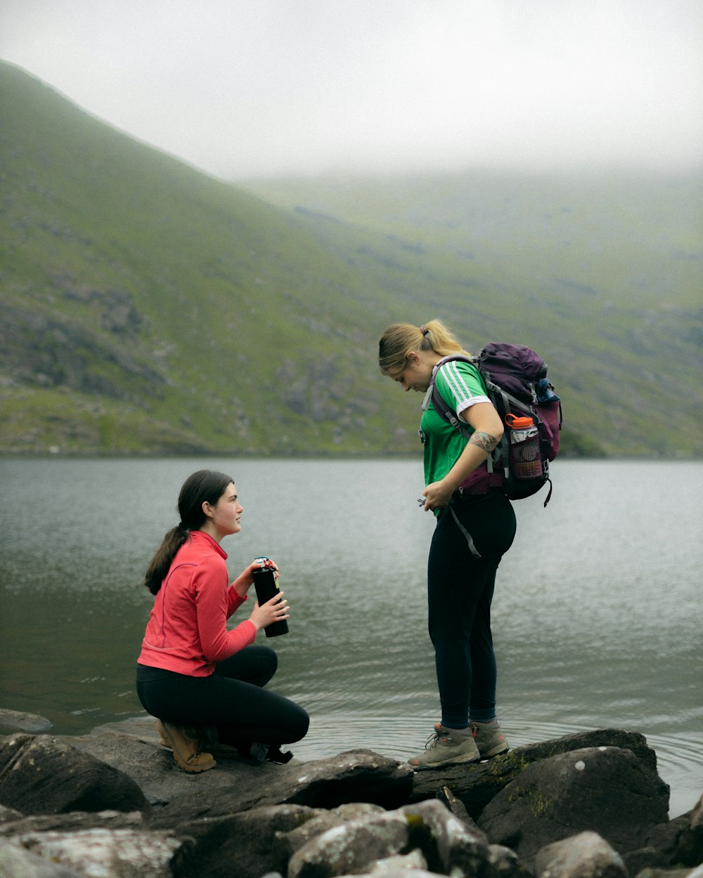 woman in red jacket and black pants carrying black backpack standing on rock by the lake