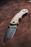 A folding knife with a textured handle and an engraved blade rests on a brown surface. The handle is crafted with a tan color and features a patterned design for grip. The blade has a matte finish with a brand name inscription near the hinge.