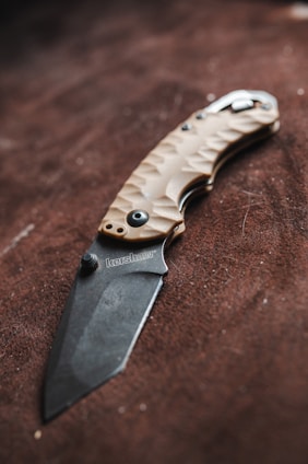 A folding knife with a textured handle and an engraved blade rests on a brown surface. The handle is crafted with a tan color and features a patterned design for grip. The blade has a matte finish with a brand name inscription near the hinge.