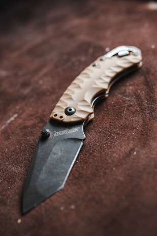 A folding knife with a textured handle and an engraved blade rests on a brown surface. The handle is crafted with a tan color and features a patterned design for grip. The blade has a matte finish with a brand name inscription near the hinge.