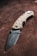A folding knife with a textured handle and an engraved blade rests on a brown surface. The handle is crafted with a tan color and features a patterned design for grip. The blade has a matte finish with a brand name inscription near the hinge.