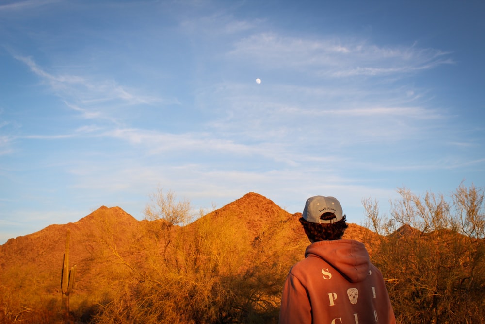 man in brown jacket wearing black cap standing near brown mountain under blue sky during daytime