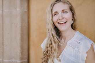 A woman with long, wavy blonde hair is smiling warmly. She is wearing a white blouse with lace details and earrings. The background features a textured, light brown wall and a stone column.