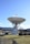 A large satellite dish antenna is positioned in a grassy area. The structure is metallic and intricate, towering over the landscape. In the background, there are rolling hills or mountains under a clear blue sky. A few trees and buildings surround the antenna.