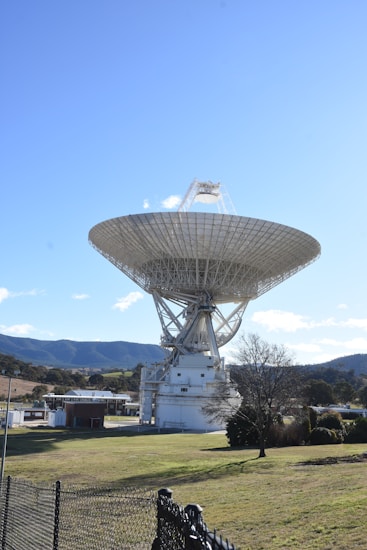 A large satellite dish antenna is positioned in a grassy area. The structure is metallic and intricate, towering over the landscape. In the background, there are rolling hills or mountains under a clear blue sky. A few trees and buildings surround the antenna.