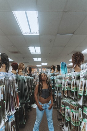 A person stands in the aisle of a store that sells hair extensions and wigs. The shelves and displays are filled with various hair products, including mannequins displaying different hairstyles and packages of synthetic hair. The ceiling has fluorescent lights, contributing to the indoor lighting ambiance.