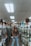 A person stands in the aisle of a store that sells hair extensions and wigs. The shelves and displays are filled with various hair products, including mannequins displaying different hairstyles and packages of synthetic hair. The ceiling has fluorescent lights, contributing to the indoor lighting ambiance.