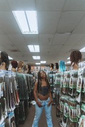 A person stands in the aisle of a store that sells hair extensions and wigs. The shelves and displays are filled with various hair products, including mannequins displaying different hairstyles and packages of synthetic hair. The ceiling has fluorescent lights, contributing to the indoor lighting ambiance.