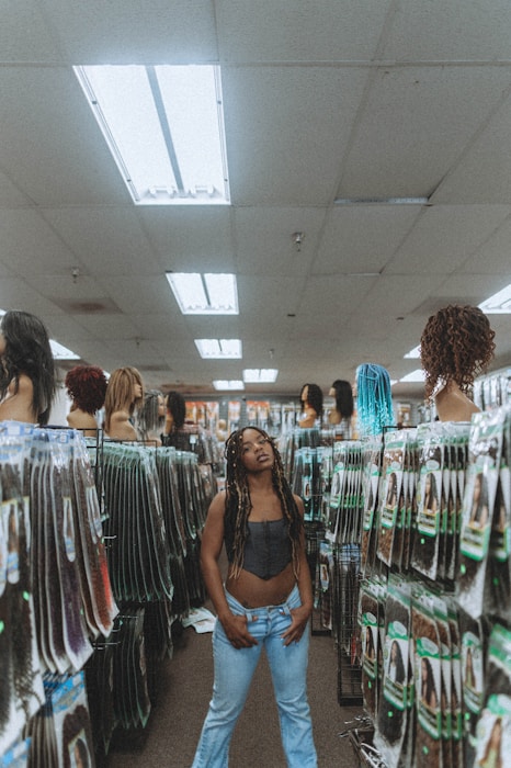 A person stands in the aisle of a store that sells hair extensions and wigs. The shelves and displays are filled with various hair products, including mannequins displaying different hairstyles and packages of synthetic hair. The ceiling has fluorescent lights, contributing to the indoor lighting ambiance.