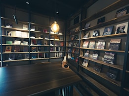 A cozy, dimly lit interior featuring wooden shelves filled with an assortment of books and magazines. A single pendant light hanging above a wooden table creates a warm ambiance. The shelves are neatly organized, showcasing a variety of book covers with different colors and designs.