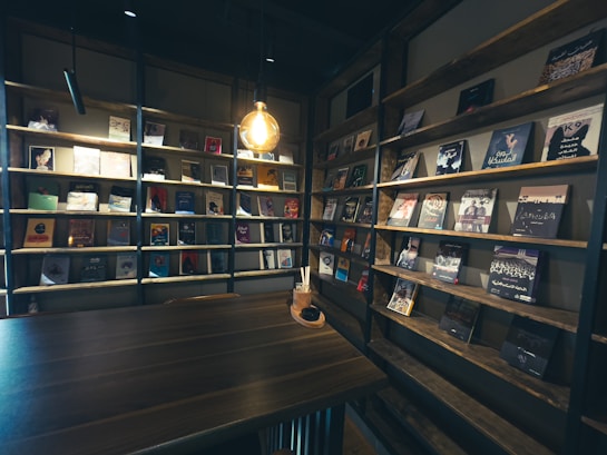 A cozy, dimly lit interior featuring wooden shelves filled with an assortment of books and magazines. A single pendant light hanging above a wooden table creates a warm ambiance. The shelves are neatly organized, showcasing a variety of book covers with different colors and designs.