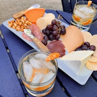 A food platter with various cheeses, crackers, grapes, nuts, and slices of salami is placed on a blue table. There are two glasses of a beverage with ice and garnished with orange slices. The arrangement suggests an outdoor dining setup, possibly for a casual gathering.
