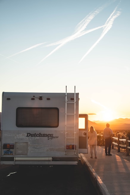 A recreational vehicle is parked on the side of the road, with two people standing nearby facing a sunset. Contrails are visible in the clear sky, adding to the serene landscape view.