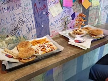A counter with a wooden surface features two trays of fast food, each containing burgers and fries. The fries on one tray are drizzled with sauces, possibly mayo and ketchup. On the wall behind, there is colorful graffiti and doodles, along with sticky notes.