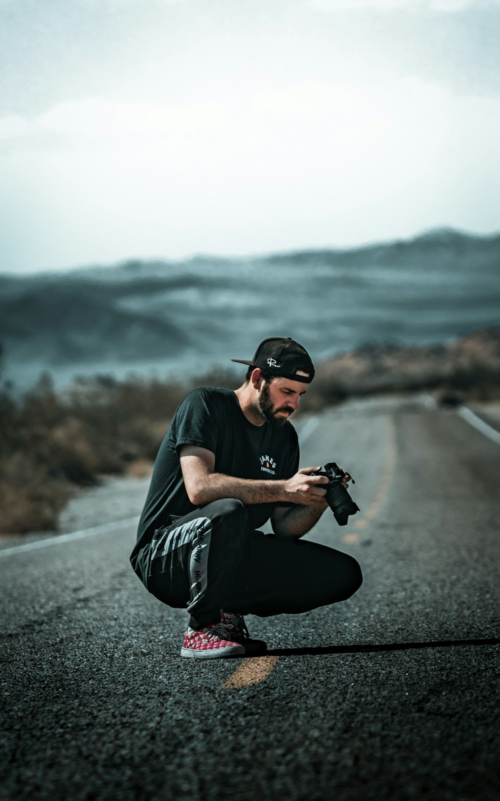 man in black t-shirt and red pants holding black dslr camera sitting on gray asphalt