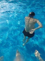 A person is swimming underwater, wearing a swim cap and shorts in a clear blue swimming pool. Sunlight reflects on the water's surface, creating shimmering patterns. There are also two feet visible below the surface, likely belonging to another person standing in the pool.
